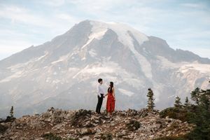 Hikers traverse a rocky ridge with majestic snow-capped Mount Rainier looming in the background of the Washington landscape.