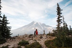 Two hikers stand silhouetted on a rocky slope with snow-capped Mount Rainier rising majestically in the background.