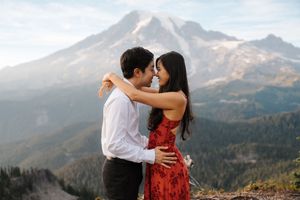 A romantic couple embraces while standing on a mountain overlook with a majestic snow-capped peak in the background.
