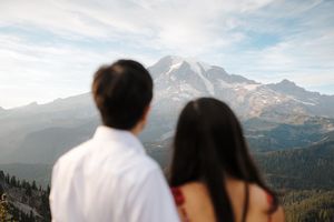 A couple looks out over a scenic mountain vista with snow-capped peaks and evergreen forests in the background.