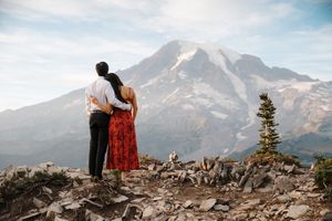 A couple stands together on a mountain peak overlooking scenic Mount Rainier in Washington State at sunset.