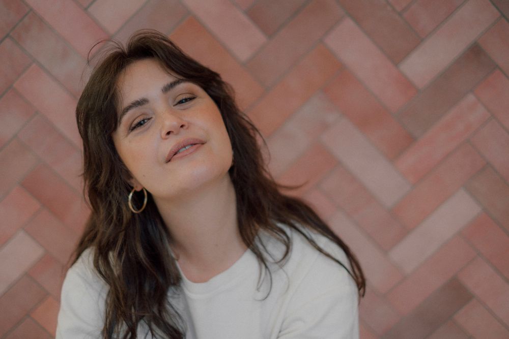 A person with dark wavy hair laughs joyfully in multiple candid expressions against a pink wall texture.