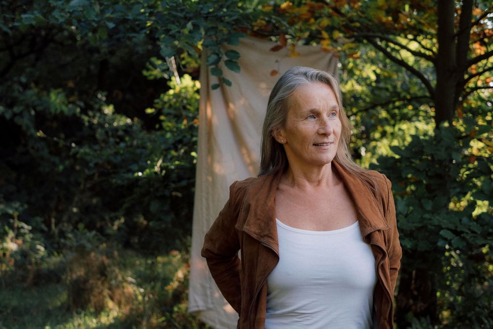 A person in a brown jacket and white top poses outdoors in a natural setting with trees and foliage.
