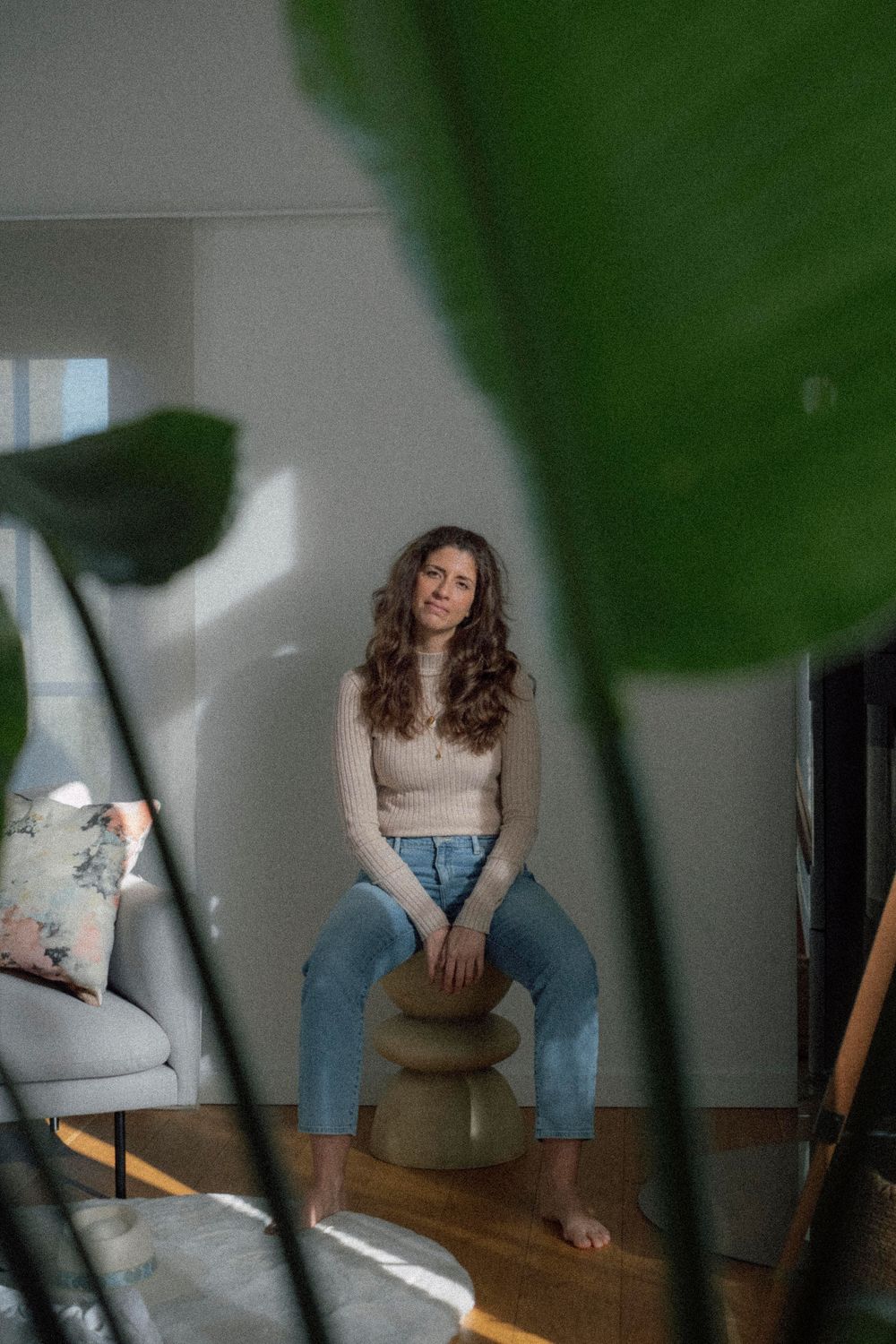 Person wearing blue jeans and beige top sits casually in sunlit room with plant shadow casting on wall.