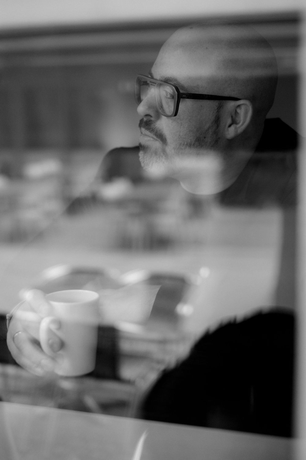 Black and white portrait through window reflection showing person with glasses holding coffee cup in contemplative moment.