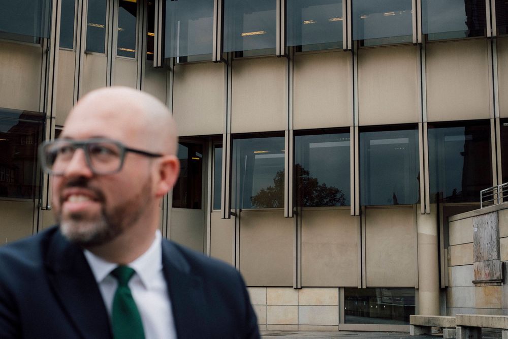 A person in a navy suit and green tie stands in front of a modern office building with reflective glass windows.