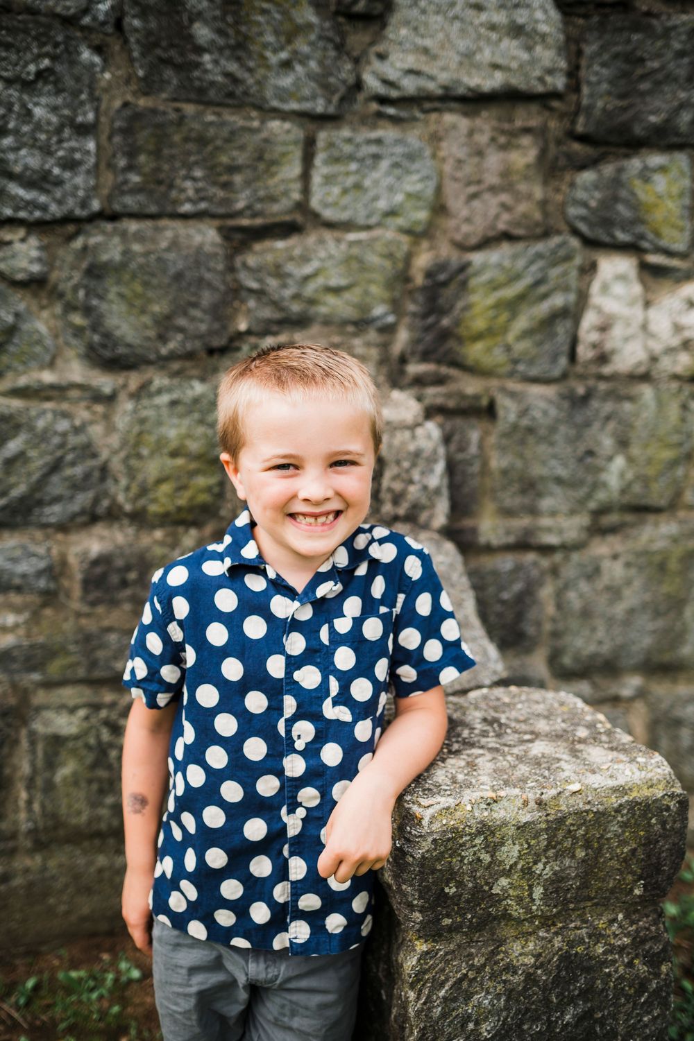 A joyful child wearing a navy polka dot shirt stands against a rustic stone wall.