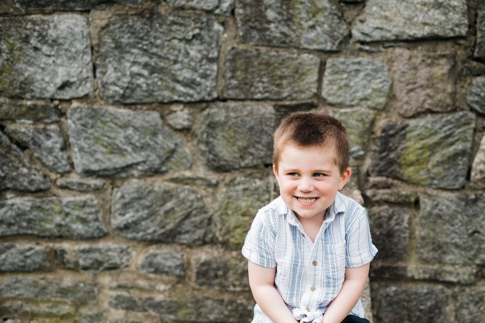 A young child in a striped shirt sits outdoors in front of a stone wall with a cheerful expression.