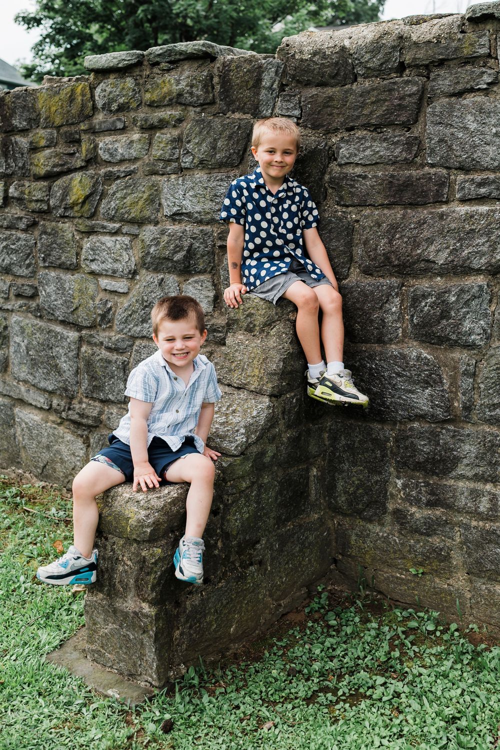 Two children sit on a rustic stone wall surrounded by grass on a summer day.