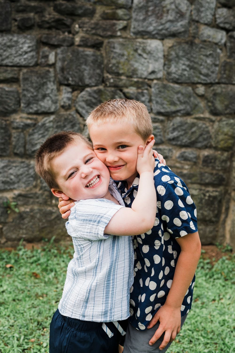 Two children share affectionate embrace and big smiles while playing outdoors against stone wall.