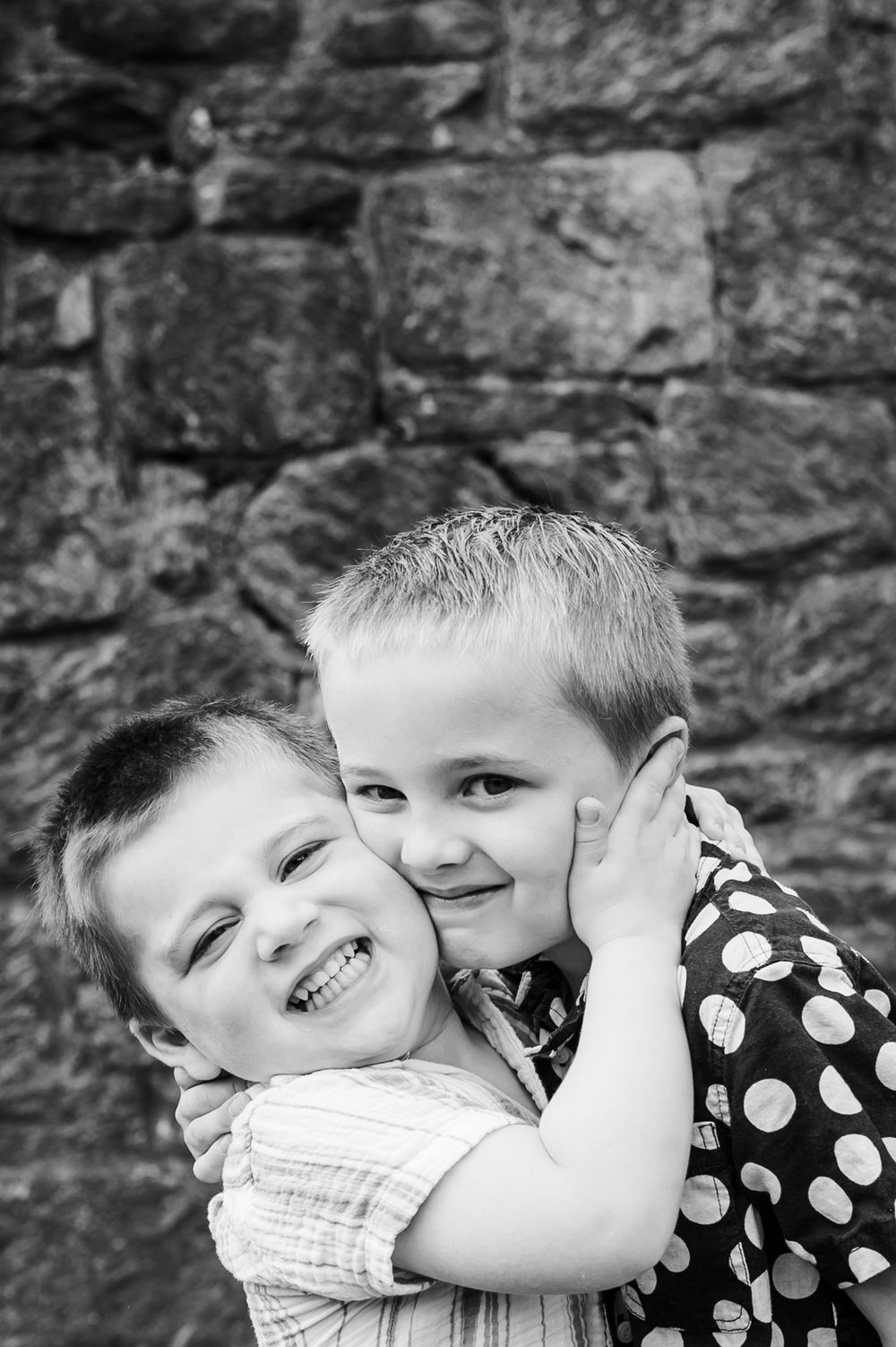 Sweet candid black and white portrait of two children hugging and laughing against a stone wall.