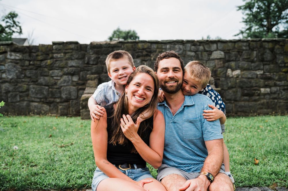 A cheerful family sits together on grass in front of an old stone wall on a sunny day.