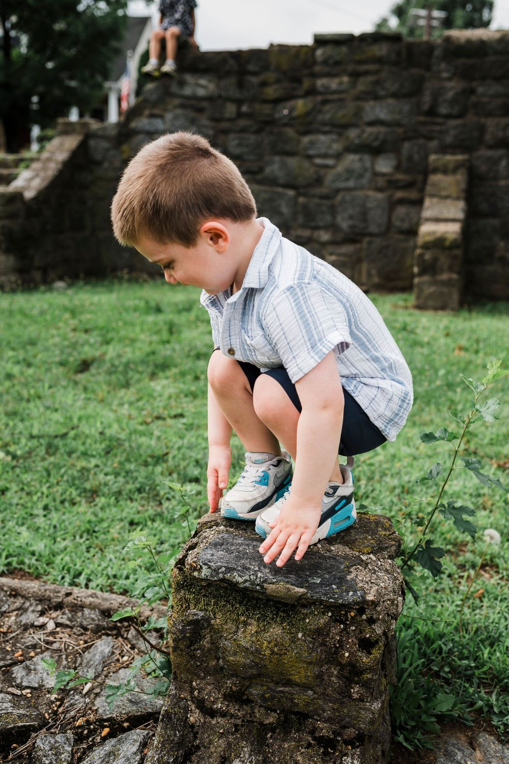 A child in casual clothes carefully examines an old stone wall while leaning over in a grassy outdoor setting.