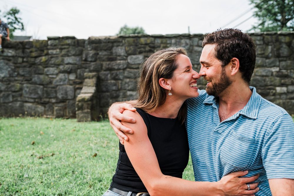 A couple shares an intimate moment while sitting together in front of a stone wall on a grassy lawn.