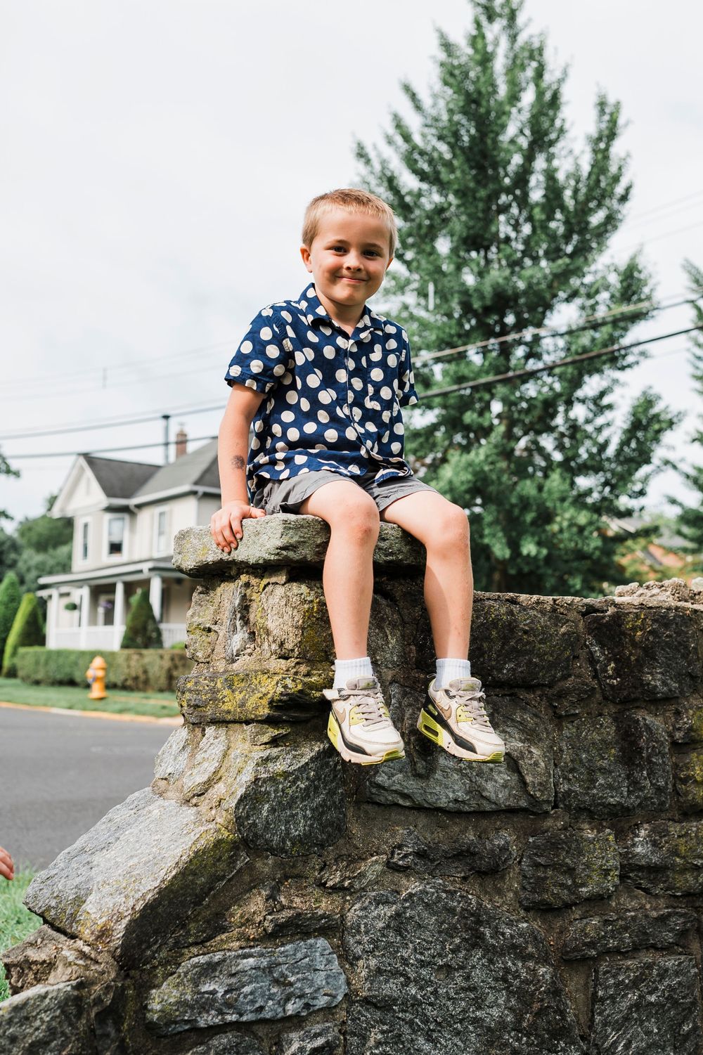 A young person in a patterned shirt sits on a stone wall in a residential neighborhood with a pine tree in the background.
