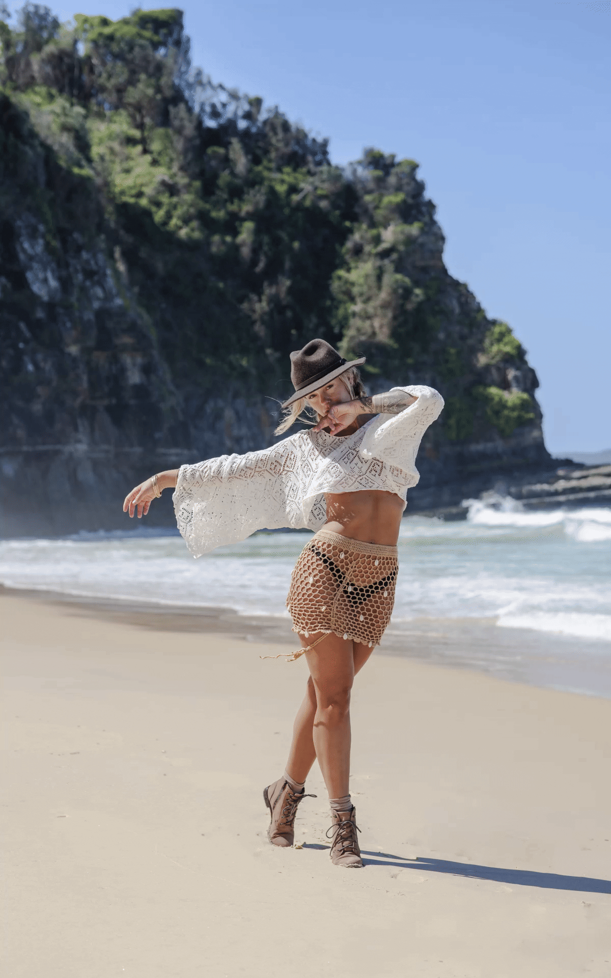 A figure in a sunhat dances gracefully on a sandy beach with a flowing white scarf against rocky coastal cliffs and ocean waves.
