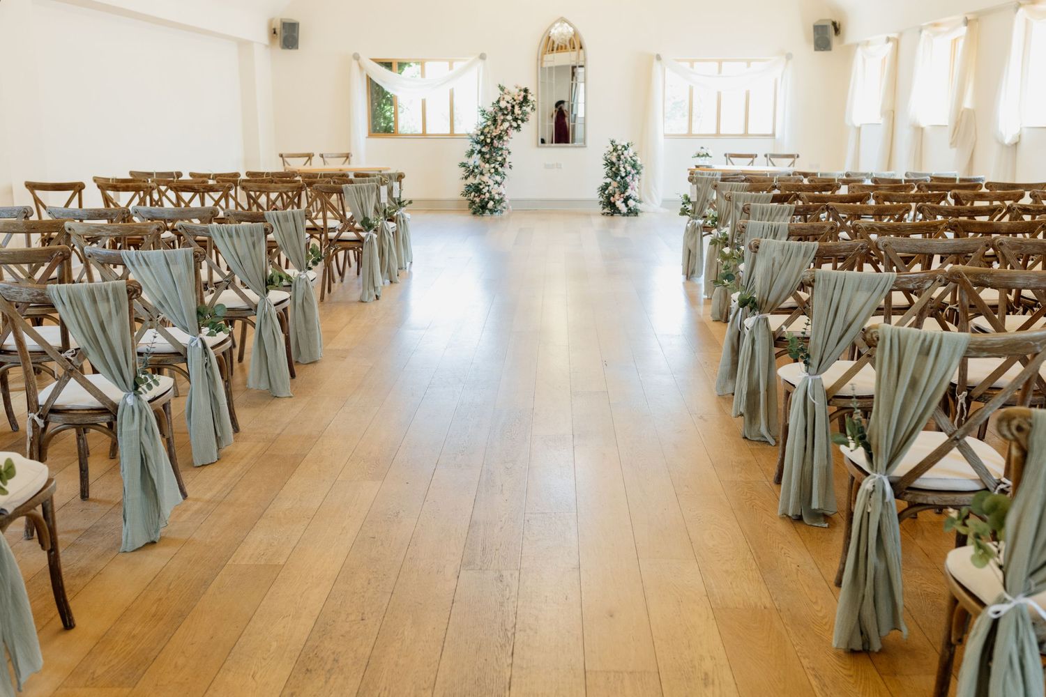 Light-filled rustic event venue with wooden cross-back chairs and white draping arranged for a ceremony.