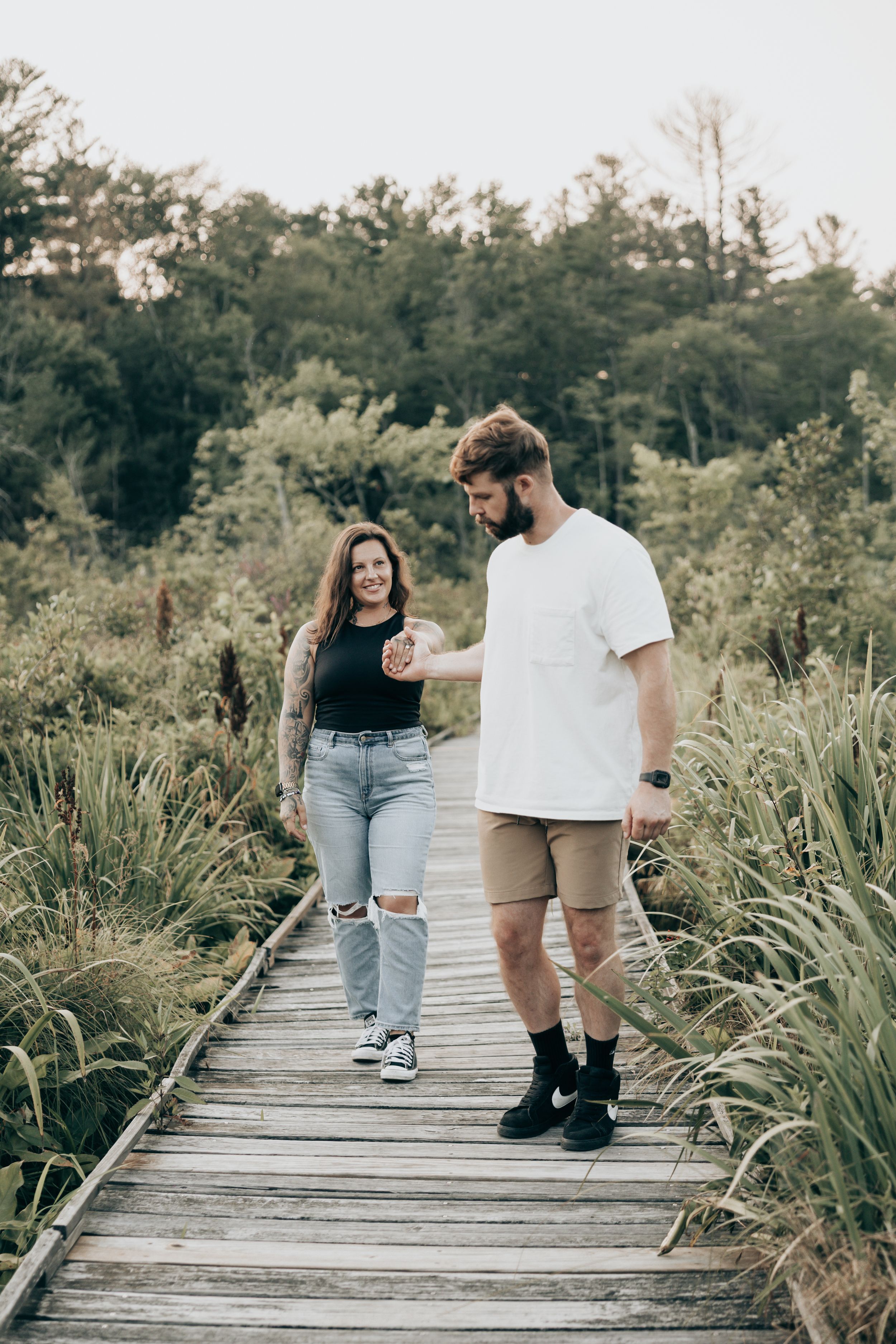 A couple walks along a wooden boardwalk surrounded by tall grass and natural foliage.