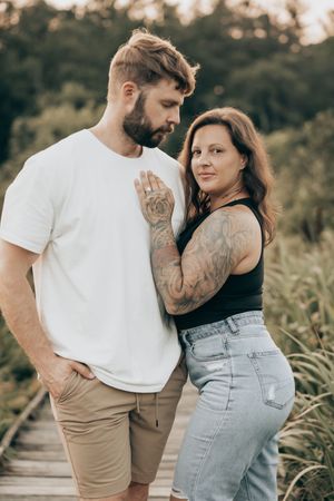 Two people embrace on a wooden path through nature, wearing casual clothing against a green background.
