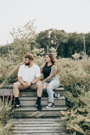 Two people sitting and talking together on wooden steps surrounded by green foliage in a natural outdoor setting.