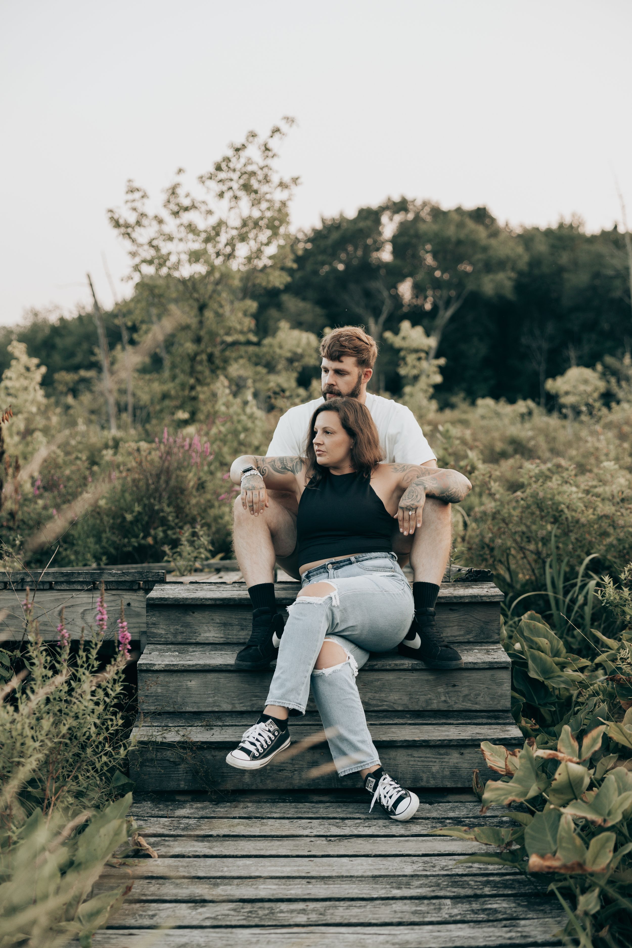 A couple sits together on a rustic wooden boardwalk surrounded by lush greenery in a casual outdoor setting.