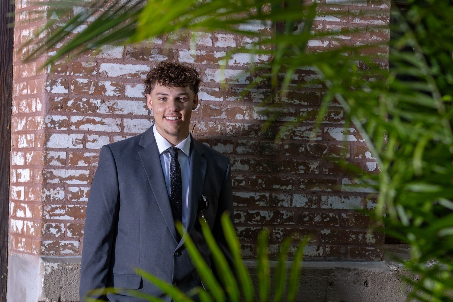 A formal portrait against a weathered brick wall with tropical plants framing the composition.