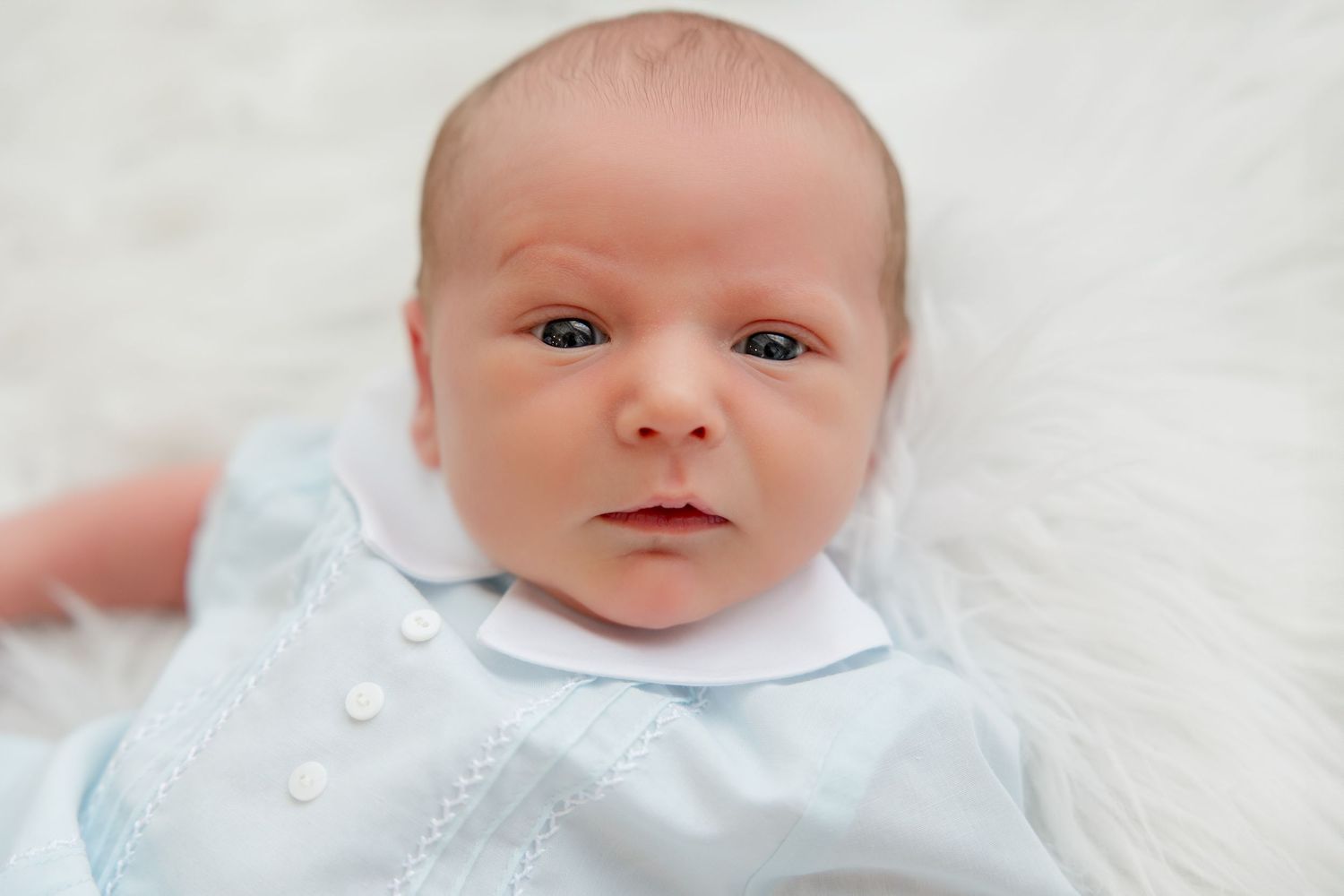 A tiny baby wearing a light blue outfit lays on white textured fabric looking directly at the camera.