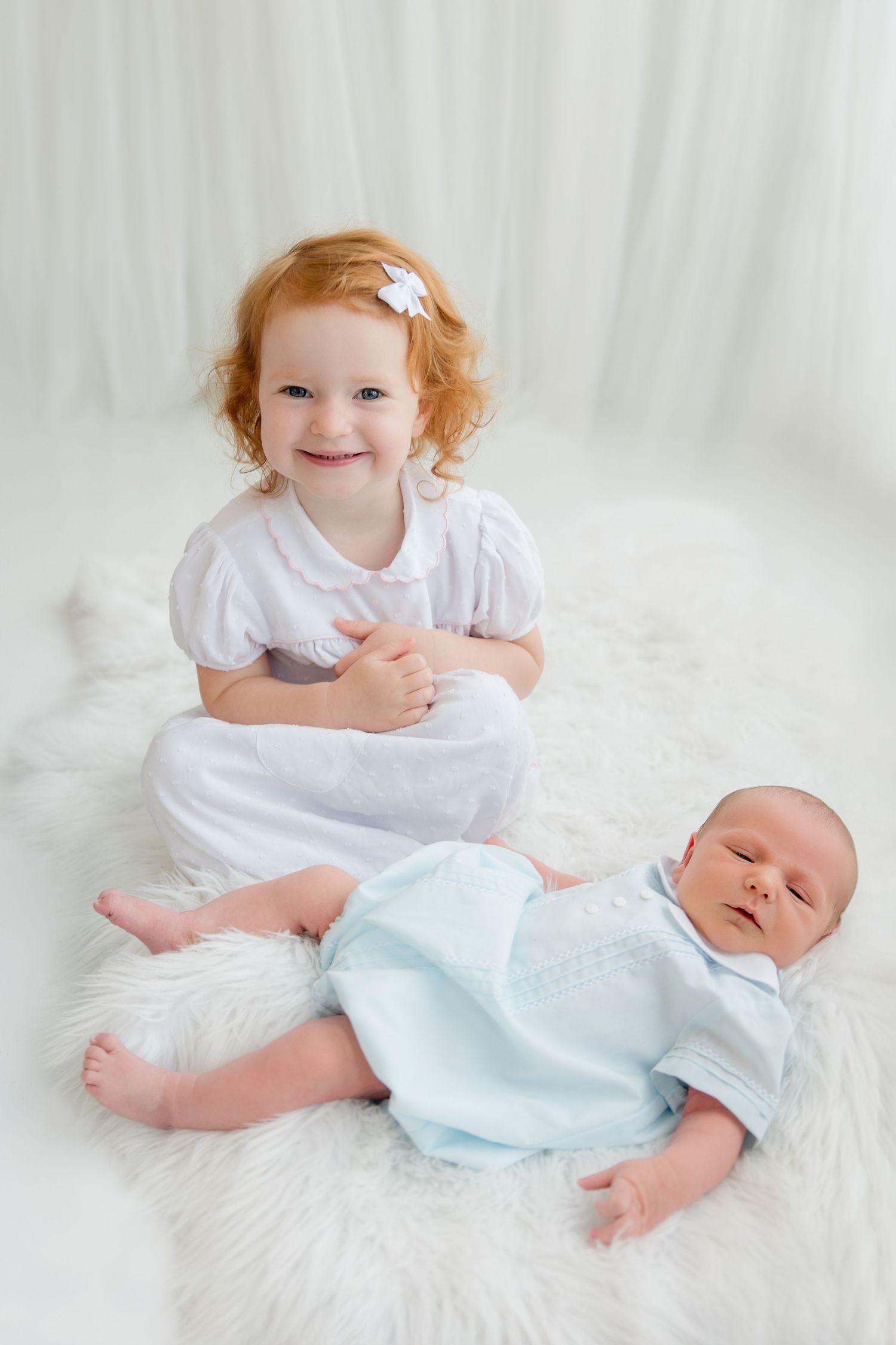 Two siblings on soft white fabric, with the older child in white dress leaning over a baby in light blue outfit.