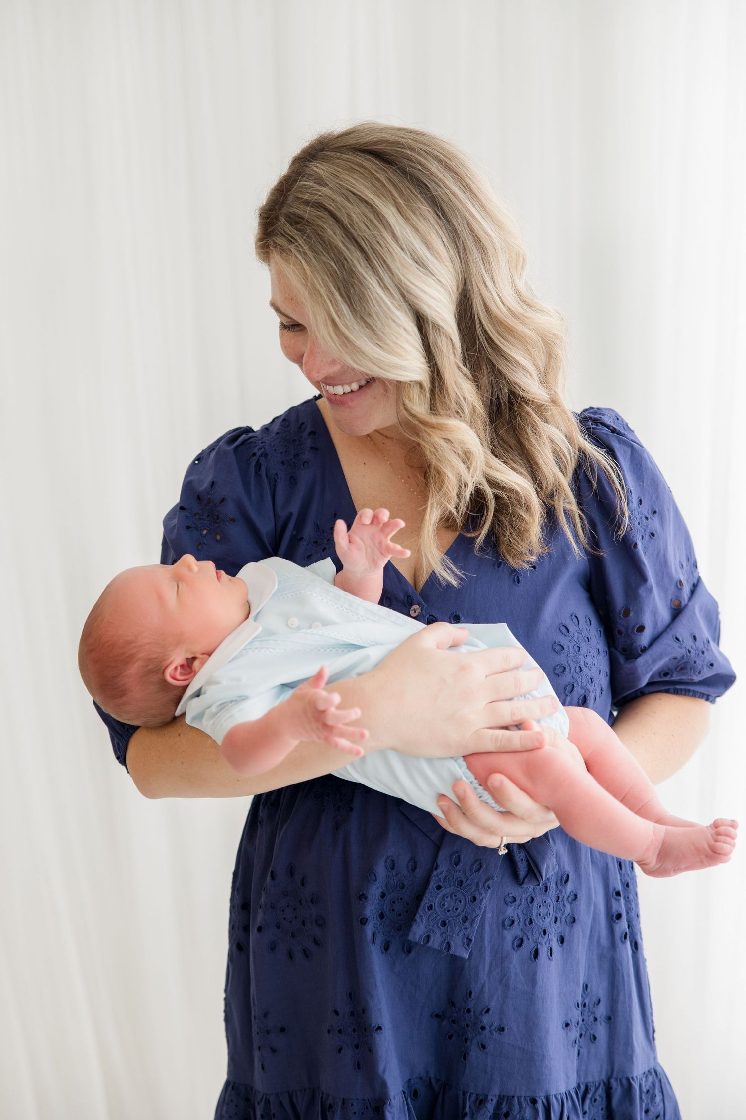 A woman in a navy blue eyelet dress holds a newborn baby while smiling and standing against a white backdrop.