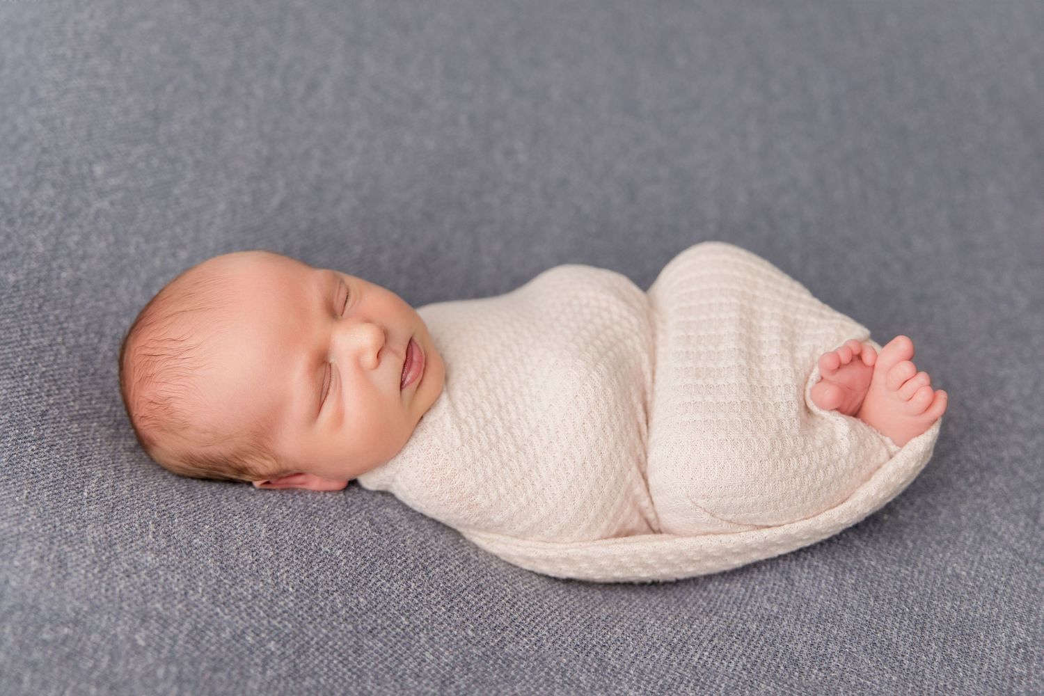 Professional studio portrait of sleeping infant posed in white wrap on textured grey background.