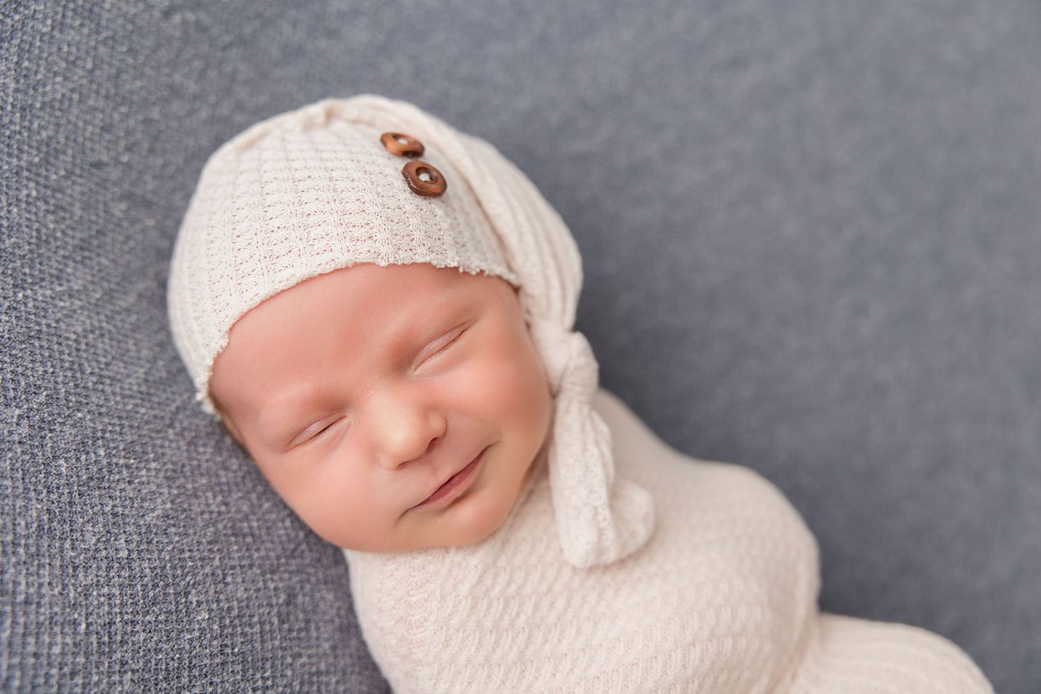A smiling newborn baby wearing a white knit cap rests on gray fabric during a professional photography session.