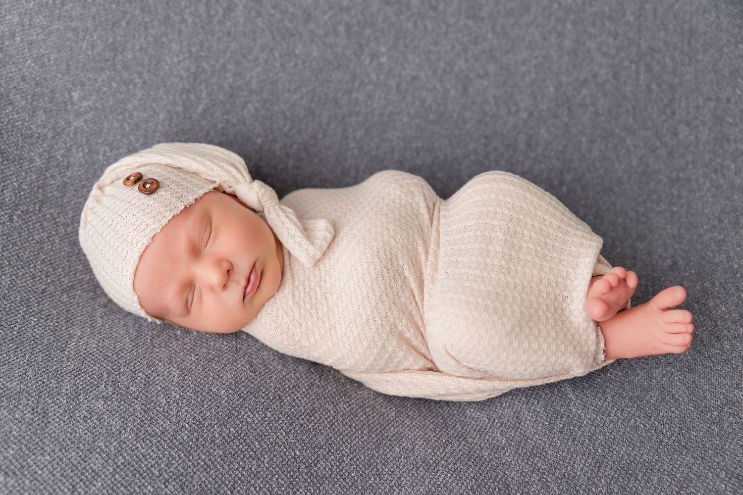 Newborn baby wearing cream colored clothing lying on grey blanket for professional photos.
