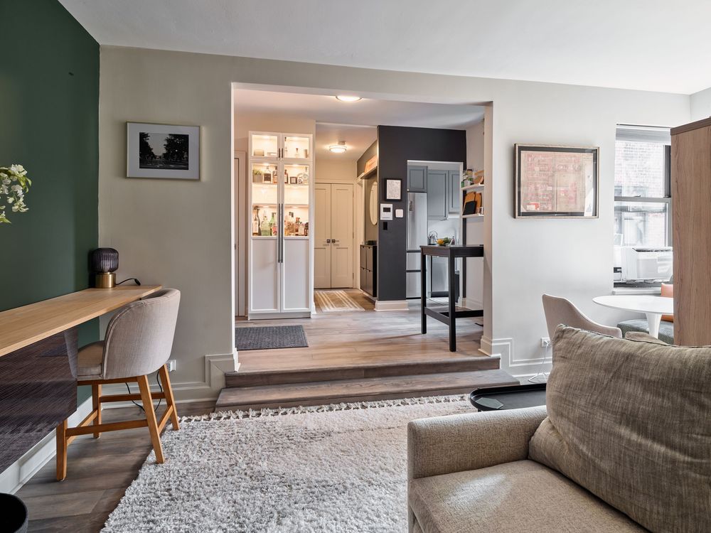 Modern living room features green accent wall, white trim, and open doorway leading to bright kitchen area.