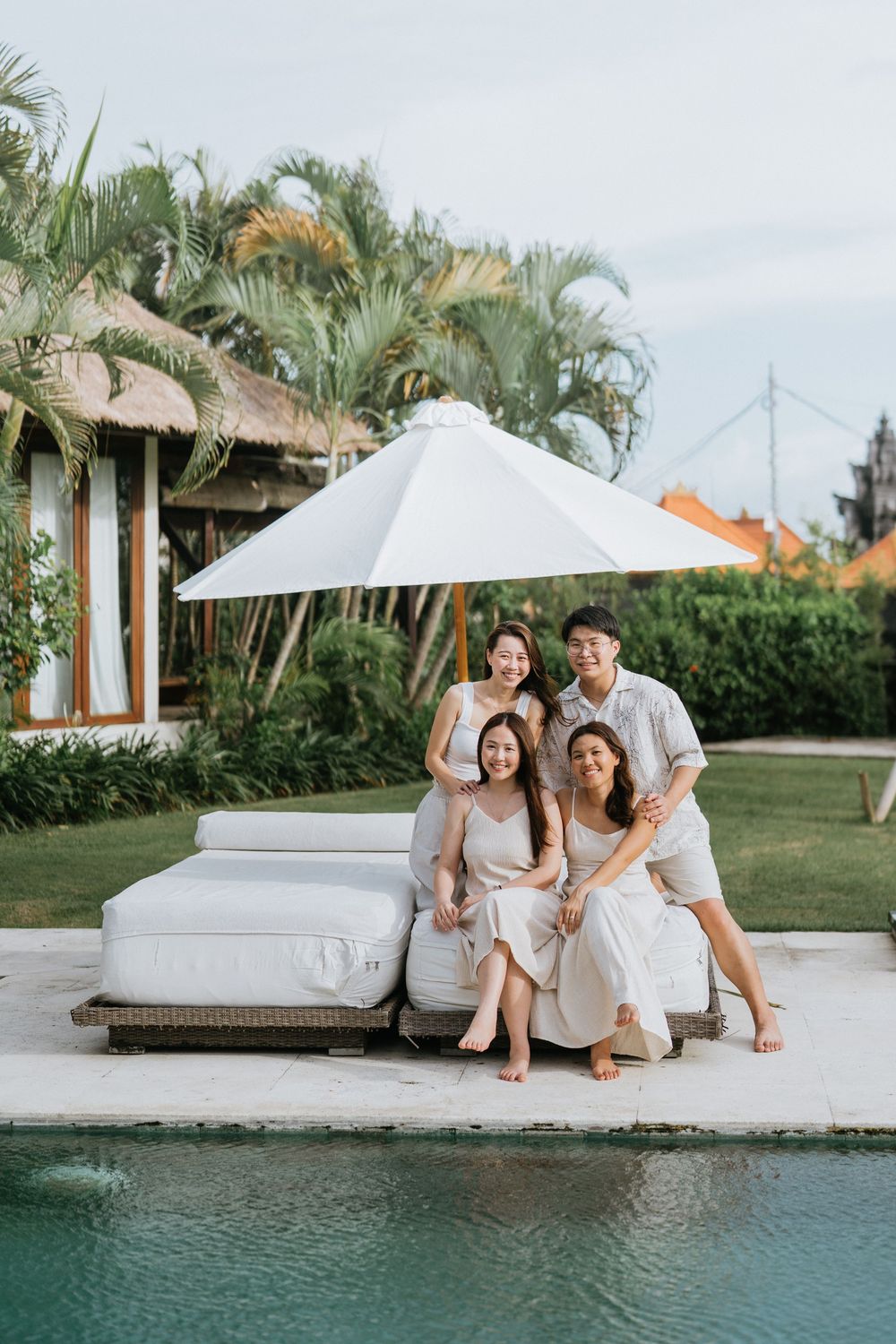 Group lounging on white outdoor furniture by a pool with tropical landscaping and thatched roof buildings.