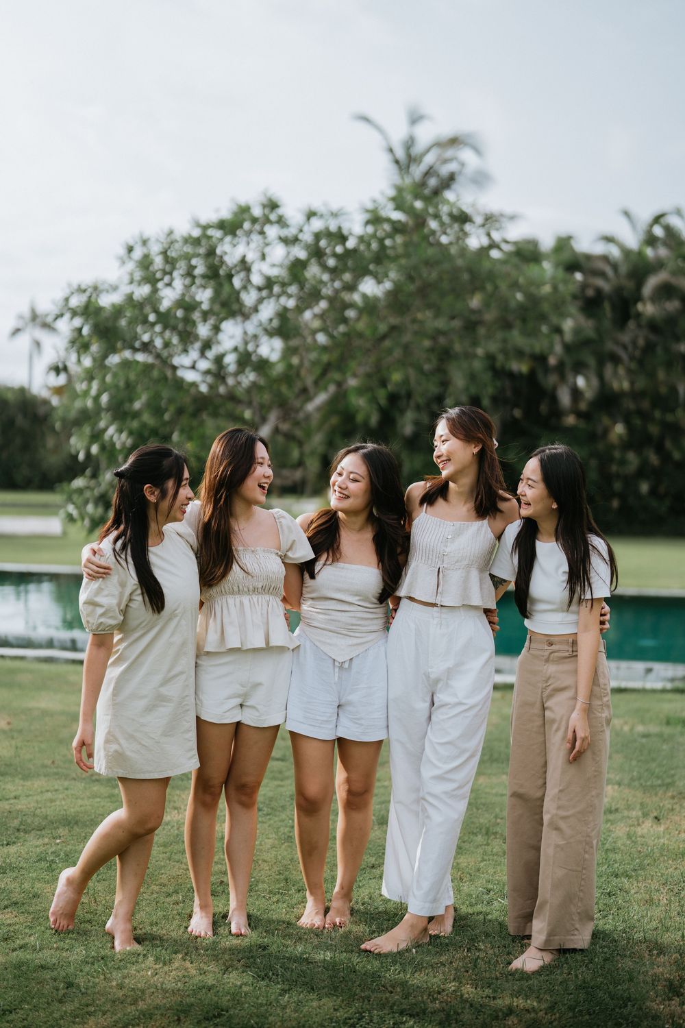 Five friends in light colored outfits laughing together on grass by a pool.