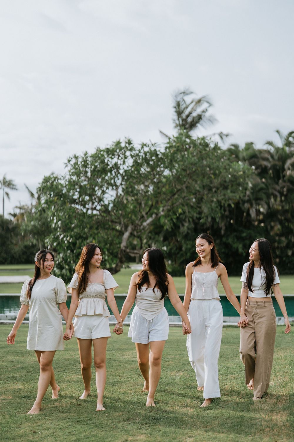 Five friends walking barefoot on grass in coordinated neutral outfits by a swimming pool.