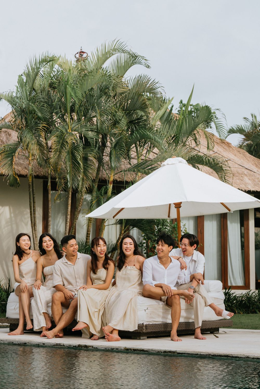 Friends lounging by tropical pool edge with palm trees and thatched roof buildings.