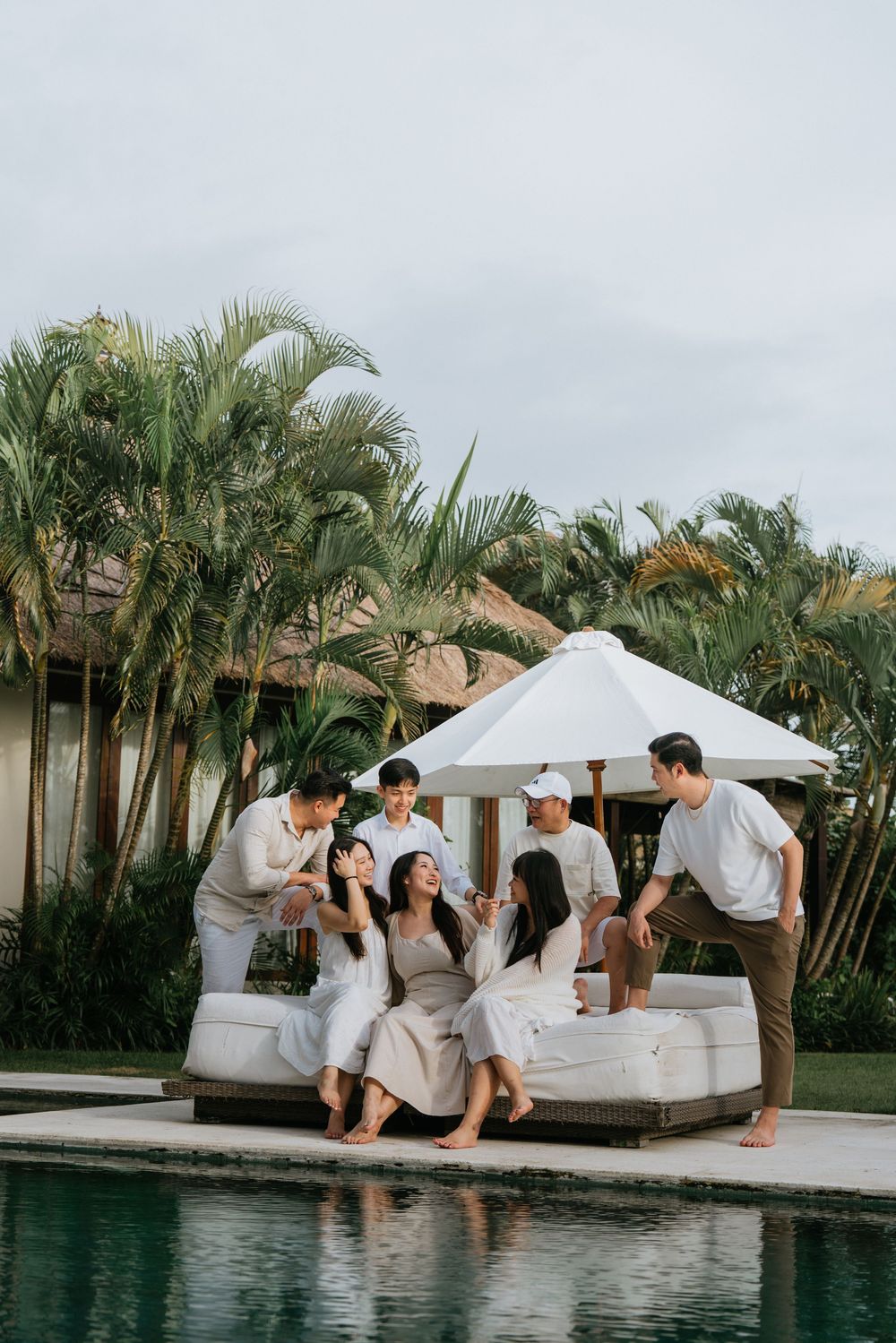 Friends relaxing on a poolside daybed with white umbrella at a tropical resort surrounded by palm trees.