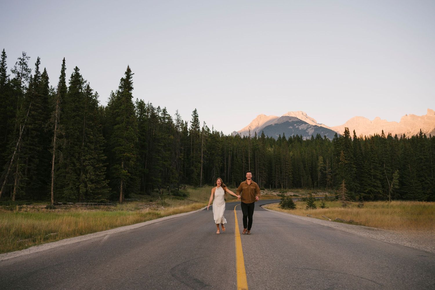 Couple running on a scenic mountain road in Banff surrounded by evergreen forest at sunset.
