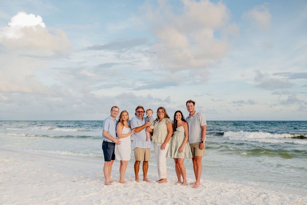 Group of people standing together on a sandy beach with ocean waves and cloudy sky in the background.