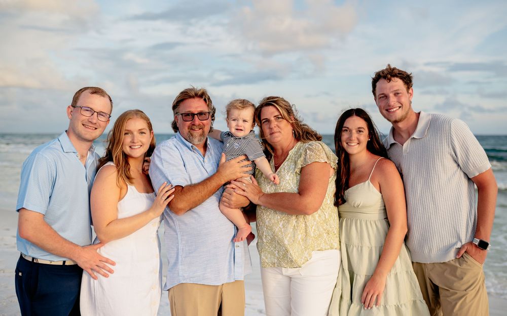 A group of people wearing coordinated white and beige outfits pose together on a beach at sunset.