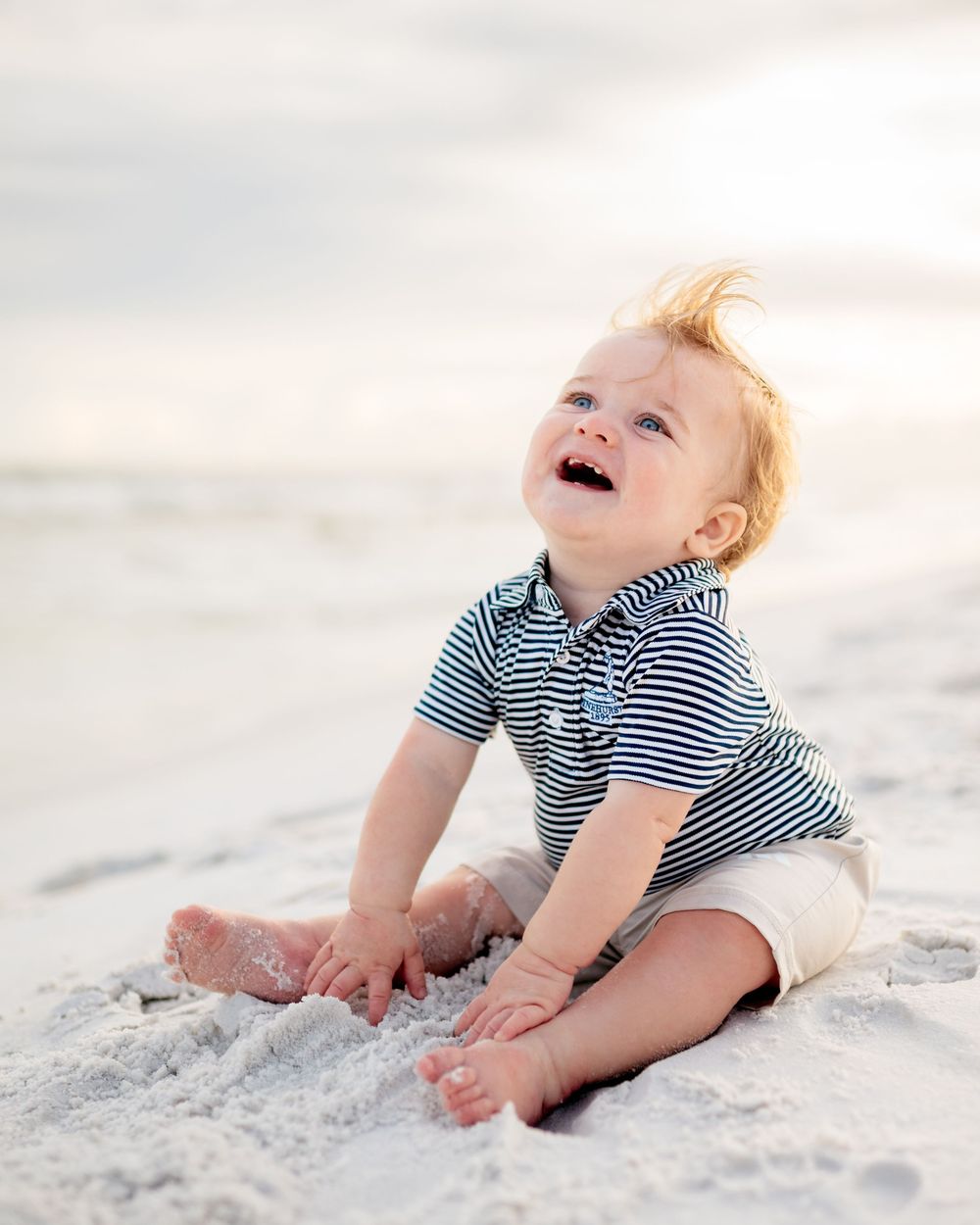 A laughing baby in a striped shirt plays on white sandy beach during a bright sunny day.