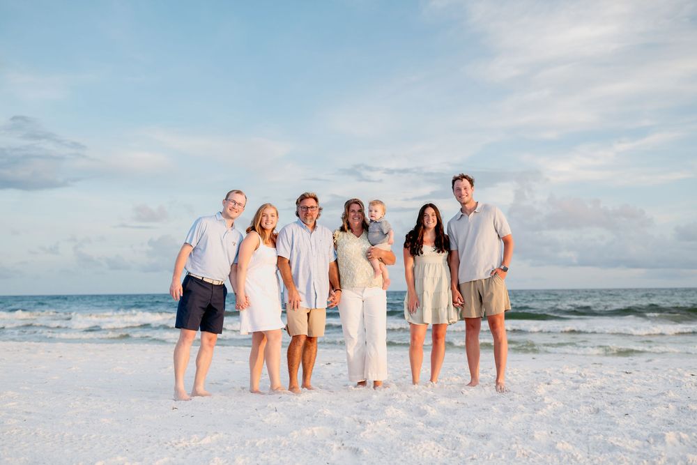 Extended family group dressed in white and blue outfits posing for photos on a scenic beach at dusk.