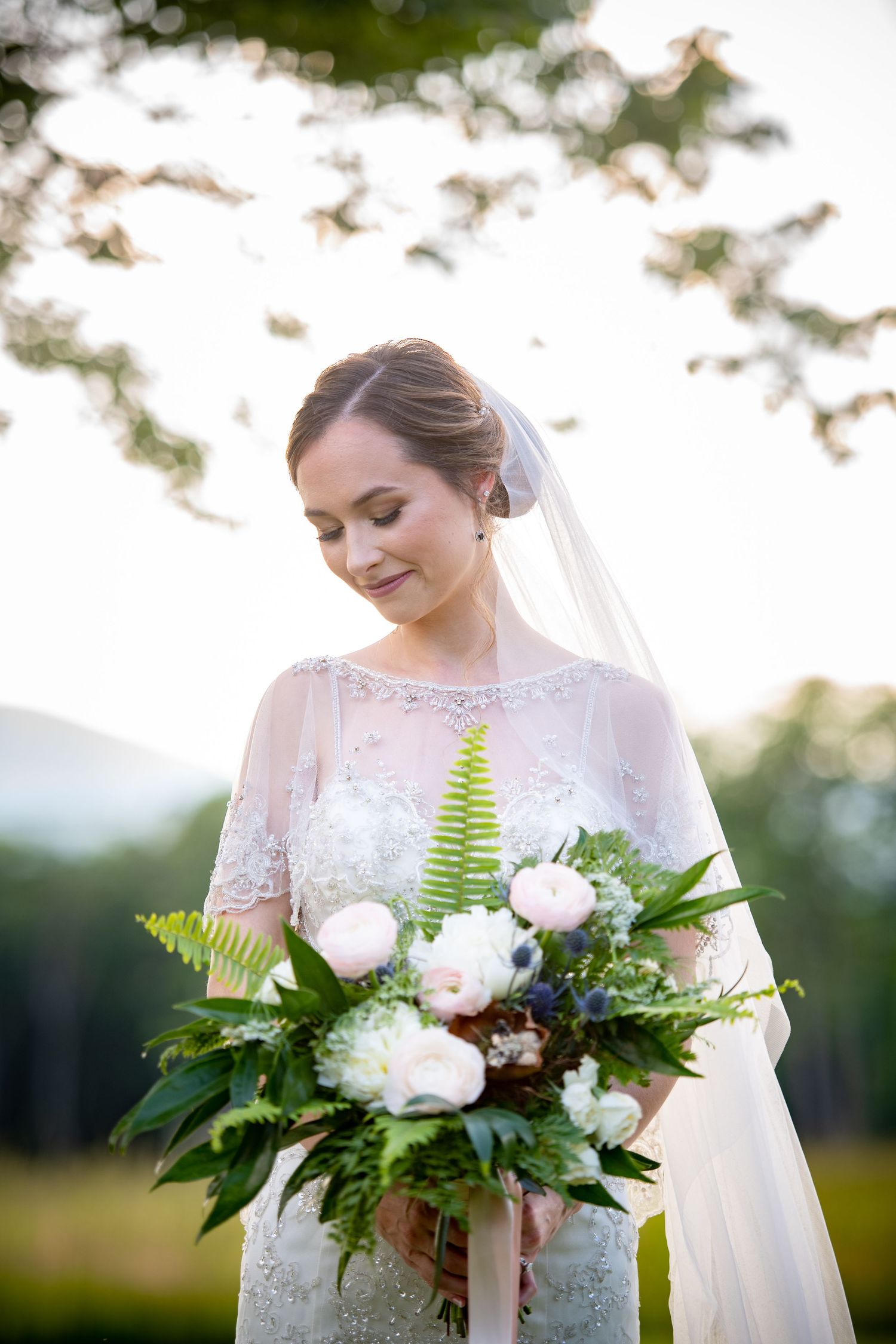 A bridal bouquet of white flowers and ferns held by someone wearing a delicate lace wedding dress at sunset.