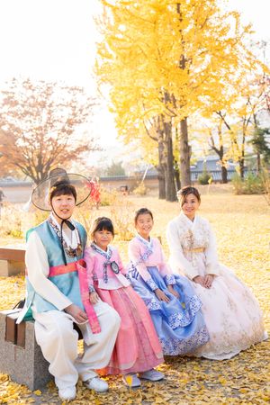 Family wearing colorful Korean hanbok traditional dress sits on stone steps among autumn leaves.