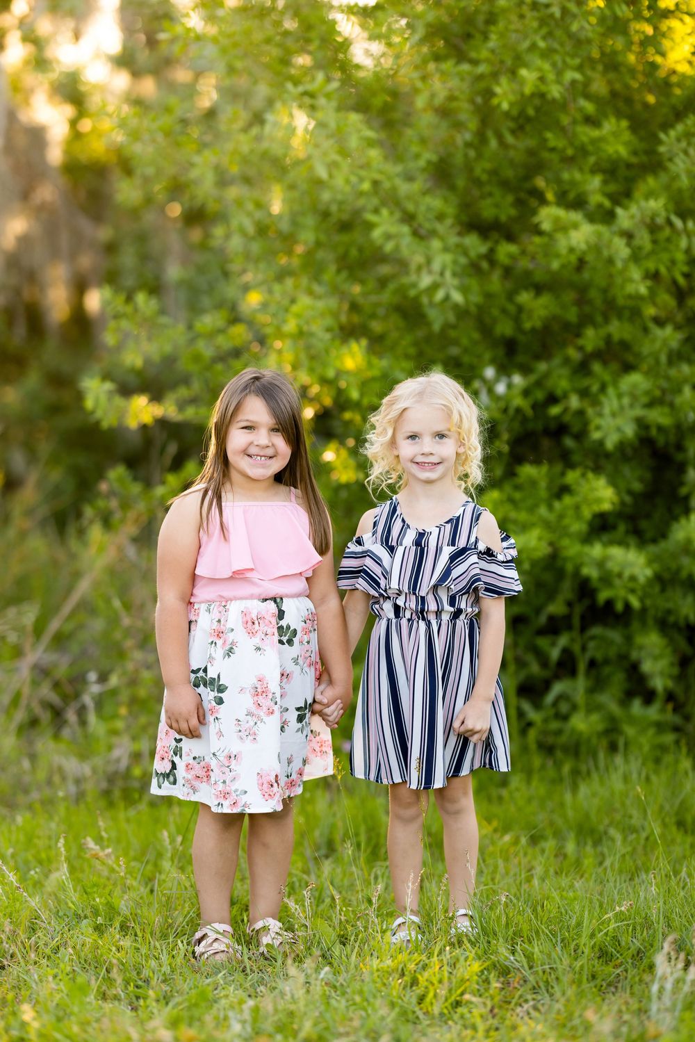 Two young children wearing pink and blue striped dresses stand together in a grassy outdoor setting on a sunny day.