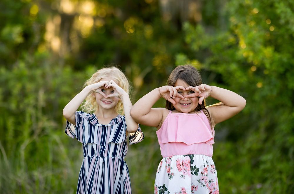 Two young children in summer dresses stand together in front of lush greenery making heart shapes with their hands.