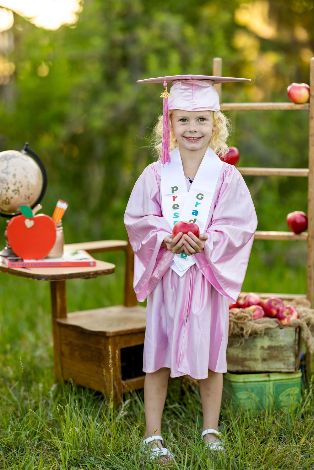 Child sitting outdoors in pink dress with wooden apple props and antique furniture for autumn photo session.