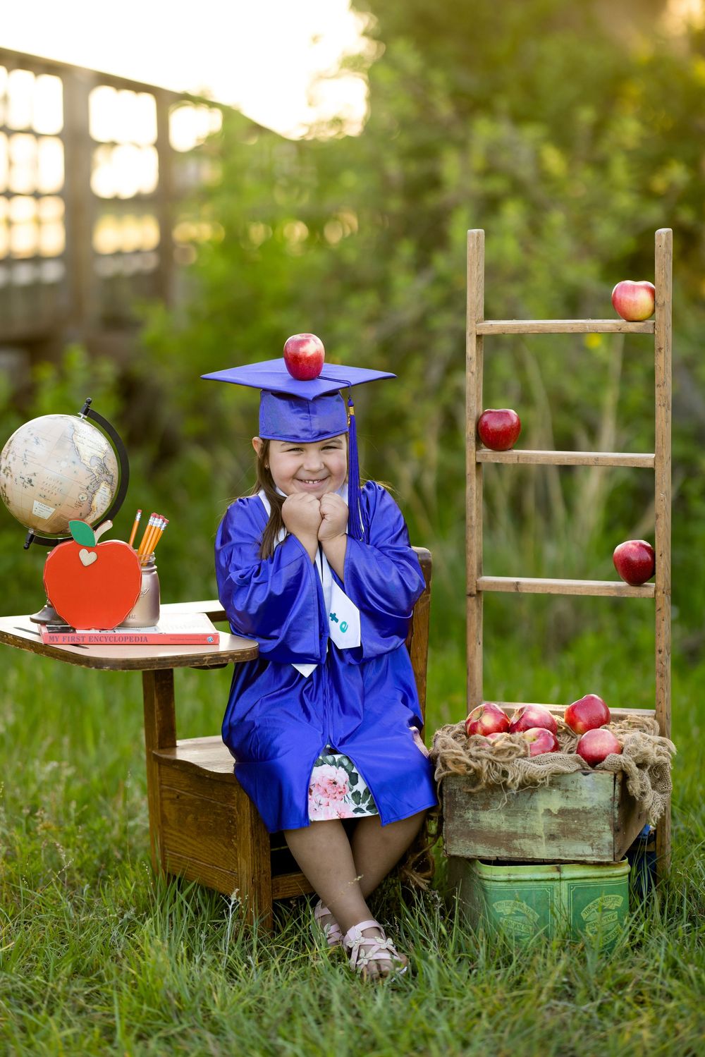 A graduation themed outdoor photo with blue cap and gown, featuring apples and school-themed decorations in a garden setting.