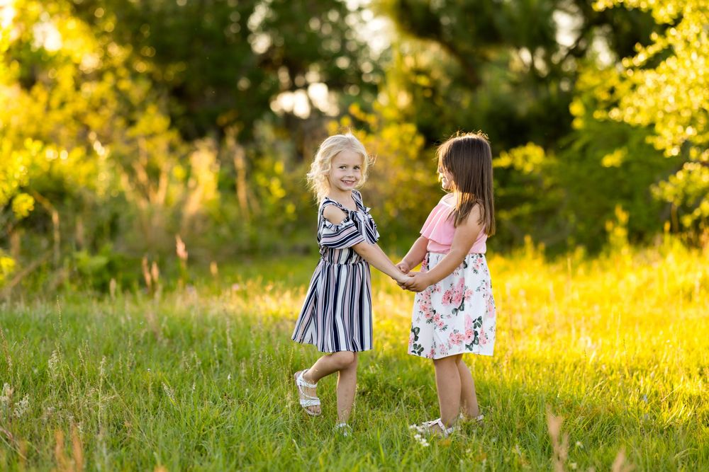 Two children in summer dresses play and laugh together in a sunlit meadow during golden hour.